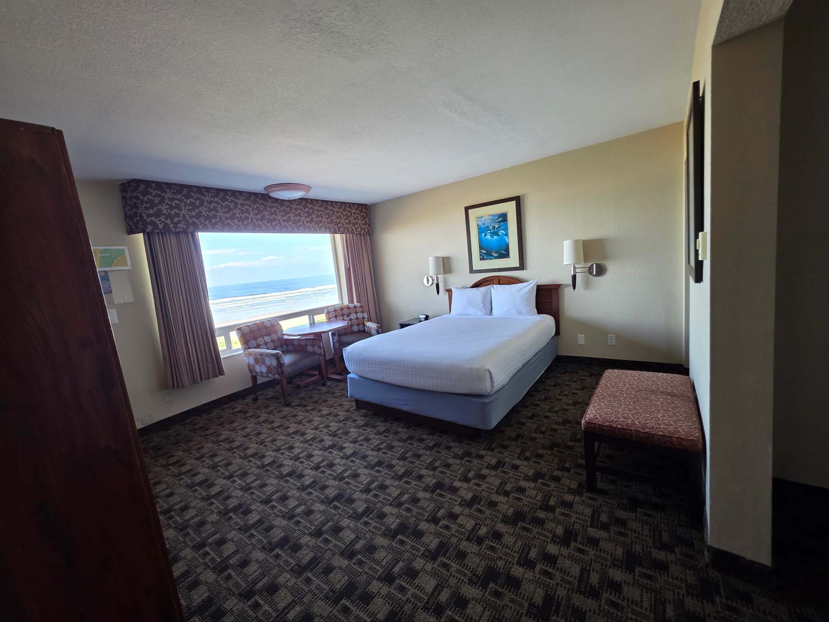 A guest room with one queen bed beside a large window with a view of the beach