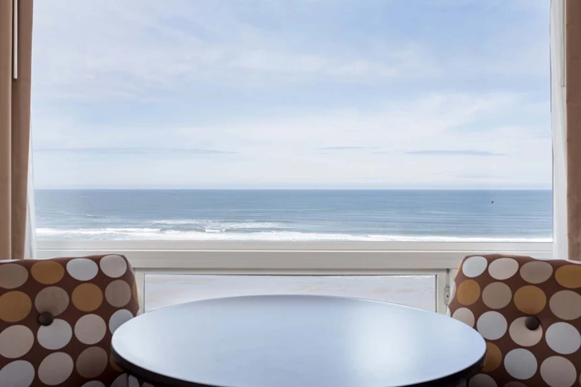 A table and two armchairs in front of a large window with a view of the beach